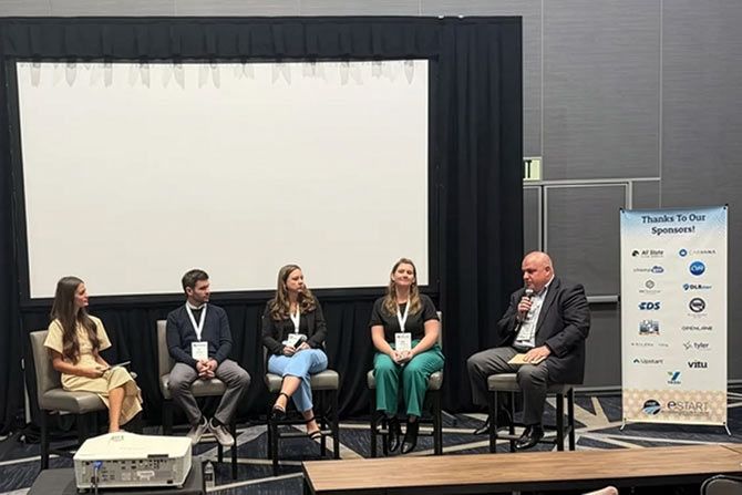 Five people are seated on a panel discussion stage with a blank screen behind them. A banner listing sponsors is to the right. The atmosphere is professional.