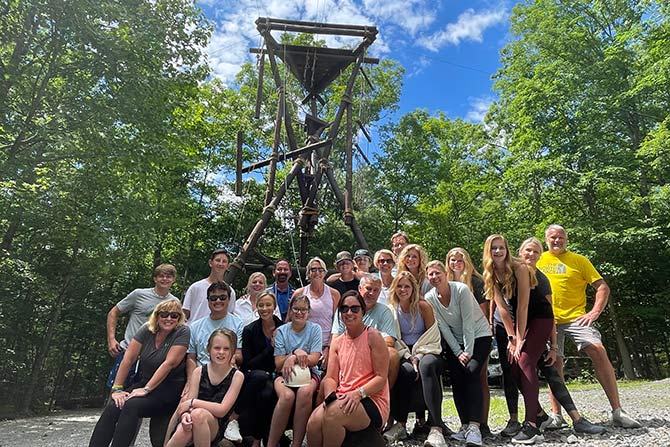 91st Annual NCADA Family Convention Returns to The Greenbrier; Group of diverse people smiling, posing outdoors in front of a wooden structure surrounded by lush green trees under a blue sky. Casual, joyful atmosphere.