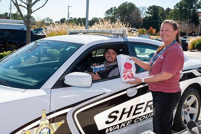 Association Happenings; A smiling sheriff in a Wake County police car receives a food package from a woman outside. The scene is friendly and the day is clear and sunny.
