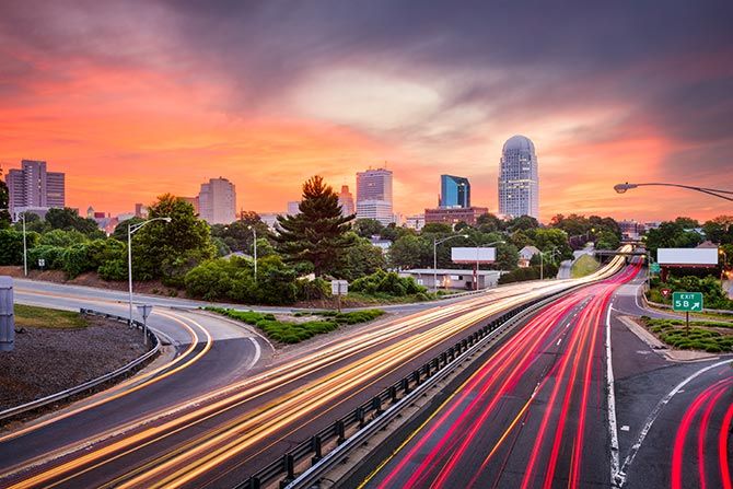 Driving North Carolina's Economy; City skyline at sunset with vibrant orange and pink skies. Blurred car lights trail on a highway, leading to tall buildings. Calm, urban ambiance.