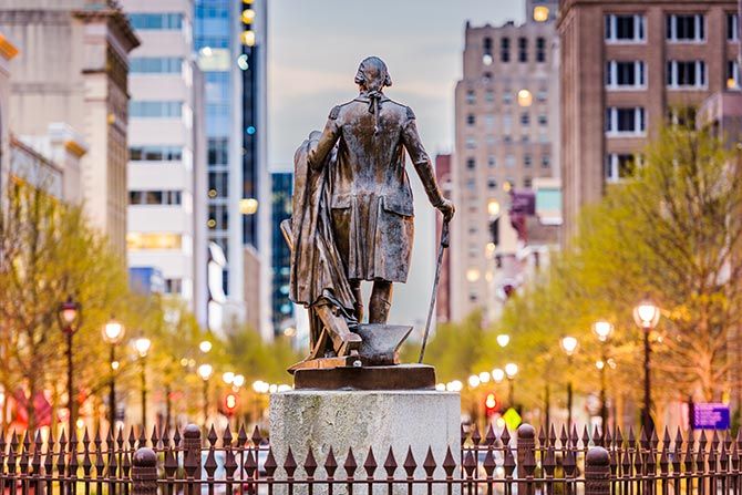 President’s Column: Leading With Unity. Bronze statue of a historical figure facing a cityscape, surrounded by trees and illuminated streetlamps, with tall buildings in the background.
