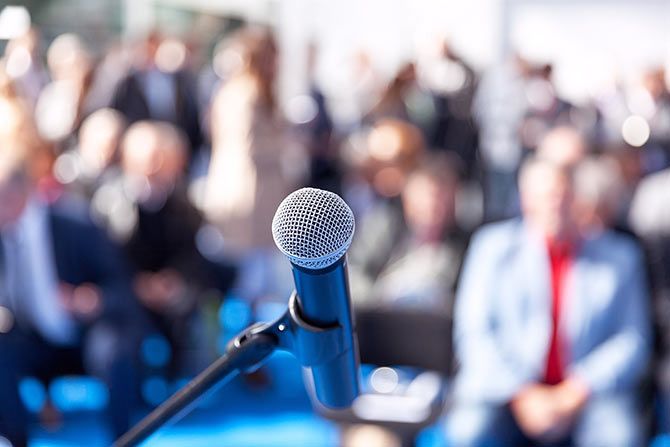 NCADA Hosts 10th Annual Controllers Seminar; A microphone on a stand in focus, set in front of a blurred crowd seated outdoors. The scene conveys anticipation and a professional event setting.