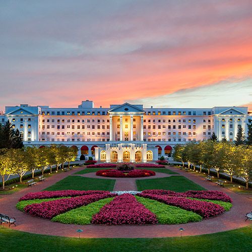 NCADA’s Annual Family Convention is Around the Corner; A grand white hotel with illuminated windows at sunset, surrounded by manicured gardens with vibrant pink flower beds and neatly lined trees.