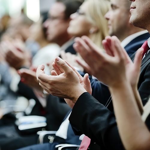 Association Happenings; Audience in formal attire clapping, focused on a presentation. The image conveys engagement and appreciation in a professional setting.