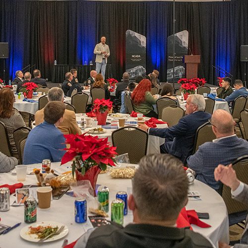 Fueling Dealers, Driving Futures; A speaker on stage addresses an attentive audience seated at round tables with red poinsettia centerpieces. The room is warmly lit, creating an engaging atmosphere.