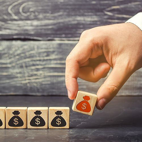 Profit Tips; A hand selects a wooden block with a red money bag and dollar sign from a row of blocks with black money bags. The scene conveys decision-making and finance.