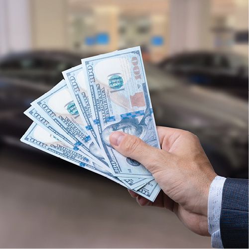 NCADA Victory Fund; A hand holding several 100-dollar bills fan-like, in focus, with blurry parked cars in the background. The scene suggests a car purchase.